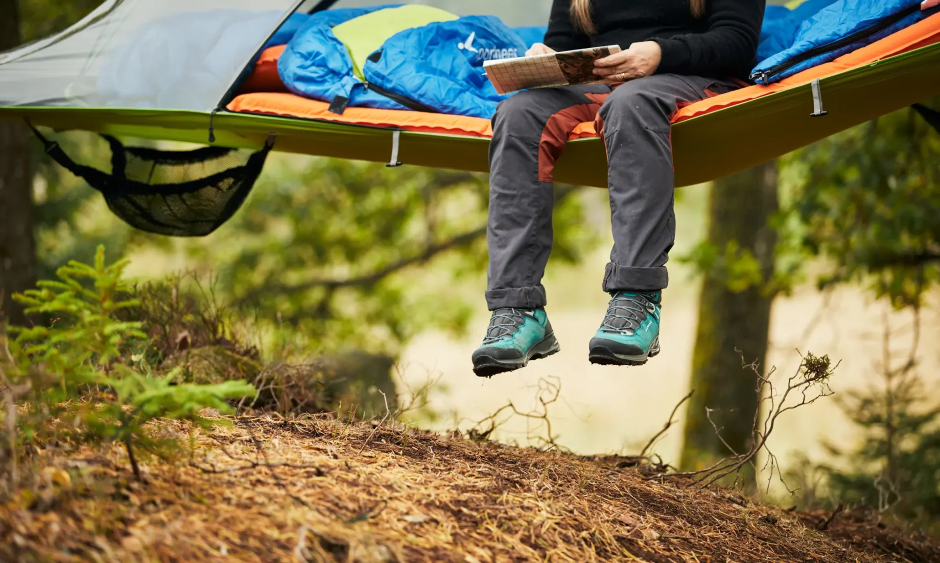 Person sitting in a suspended tree tent in the forest with hiking boots and a book in hand. Photo: Jonas Ingman.