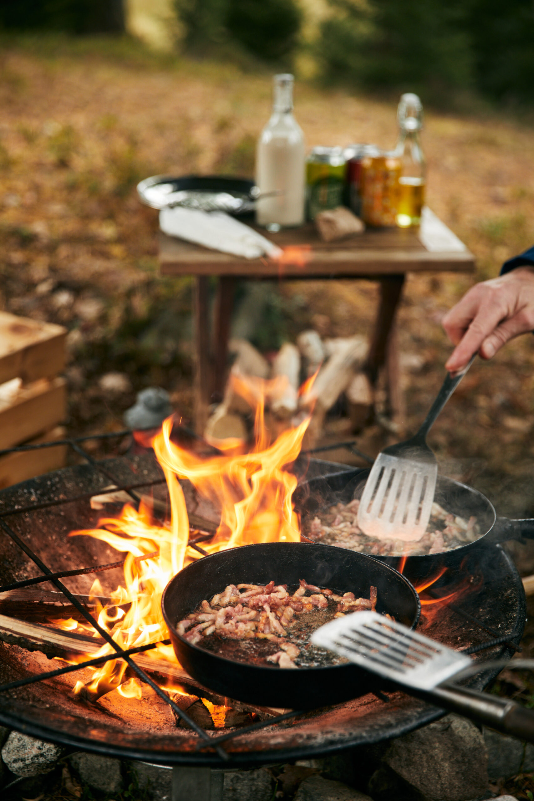 Utomhusmatlagning över öppen eld med stekpanna och höstlöv runt eldstaden.