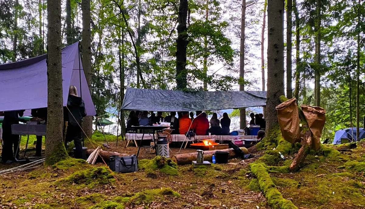 Gäster som sitter samlade under tarp i skogen vid Hjällsnäs Eco Camp, med lägereld och utrustning runtomkring, i en lugn och naturnära atmosfär.