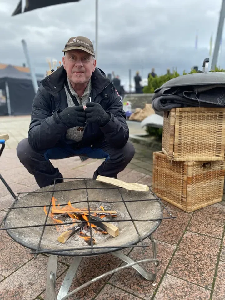 Peter Carlsson vid ett eldfat under vårmarknaden i Gråbo, omgiven av ved och friluftsutrustning.