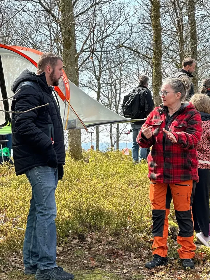 En kvinna i friluftskläder samtalar med en man under ett utomhusevenemang i skogen, med tält och deltagare i bakgrunden.