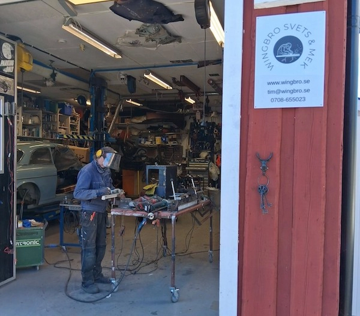 Person som arbetar med metall i en verkstad, stående i en öppen port med verktyg, maskiner och en bil i bakgrunden.