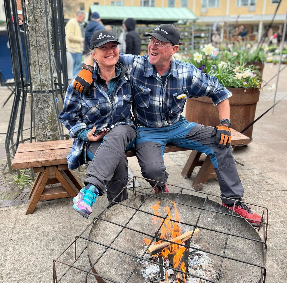 två glada personer sitter på en bänk, framför står ett eldfat där elden binner. personerna sitter på ett torg